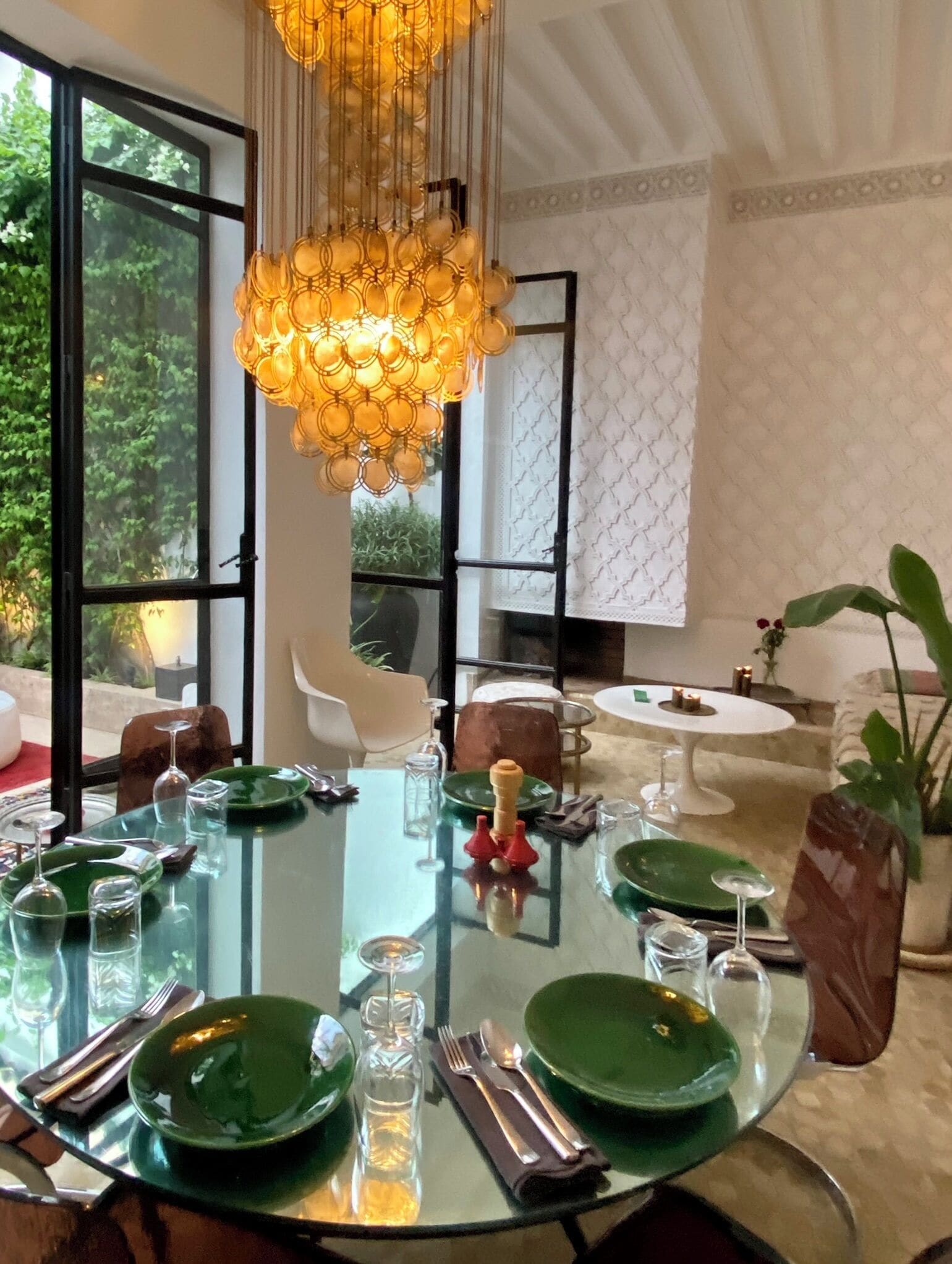 Un comedor con una mesa redonda de cristal preparada para cuatro personas, rodeada de sillas. Un candelabro cuelga sobre la mesa.