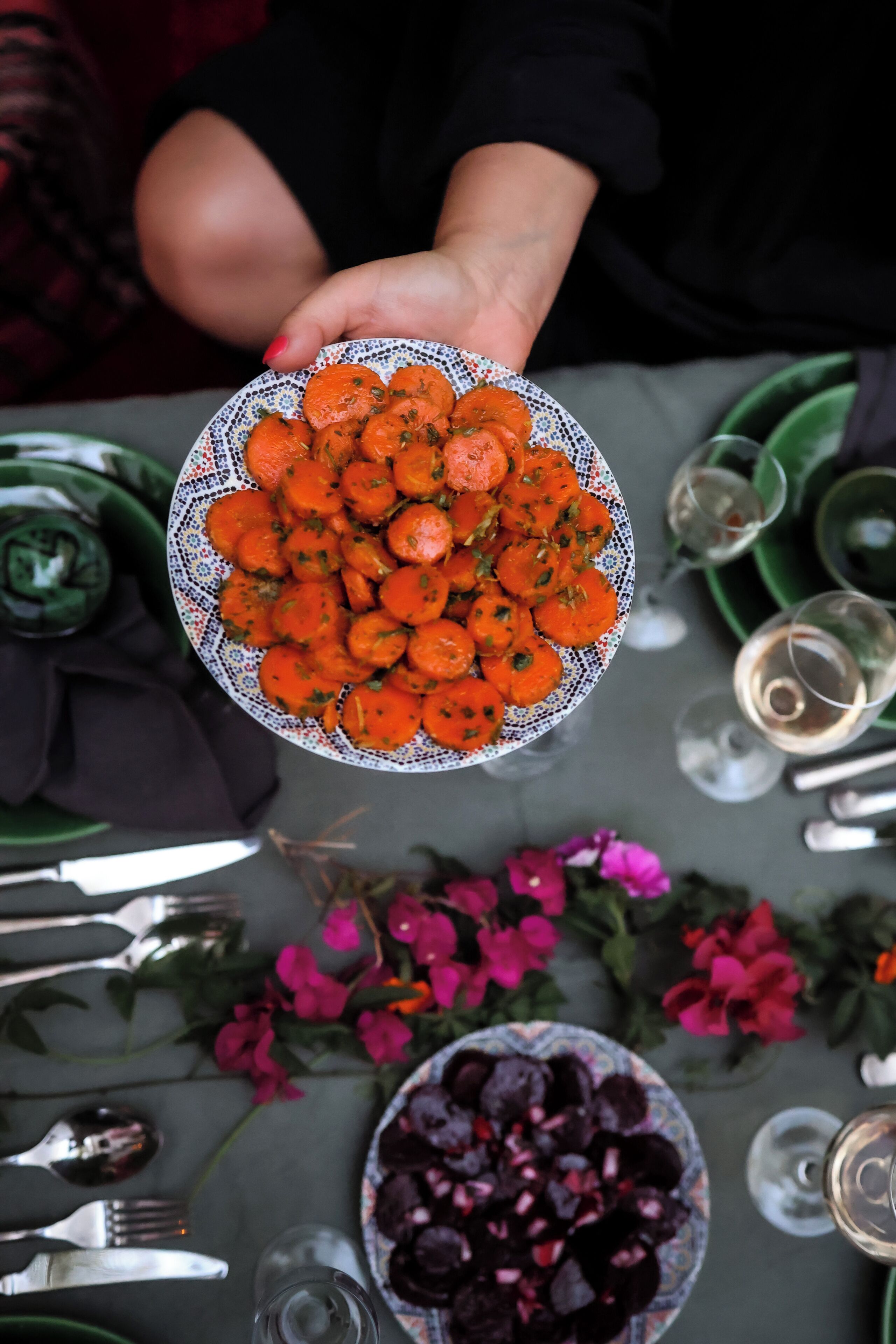 Una persona sostiene un plato con verduras anaranjadas sobre una mesa de comedor con diversos platos, utensilios y flores.