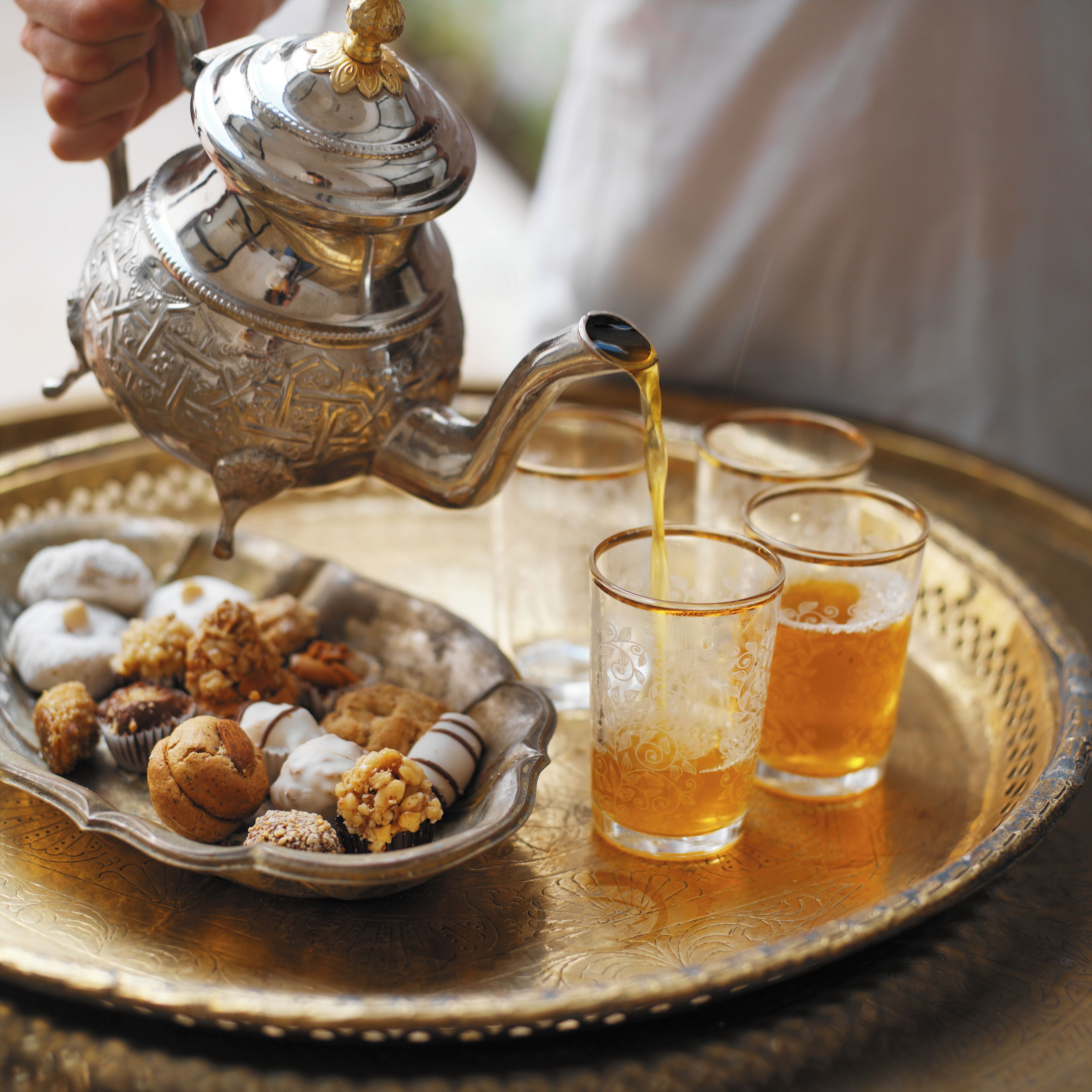 A tray with a teapot pouring tea into glasses and a plate of pastries.
