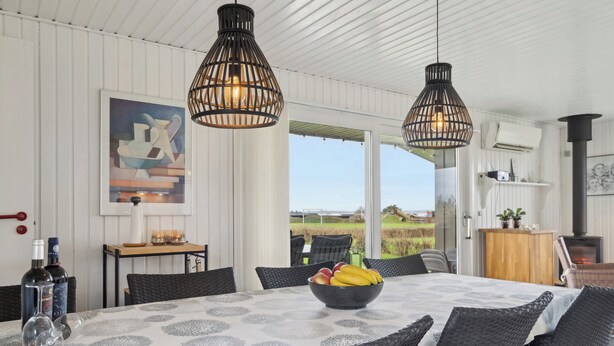 A dining area with a table set for four, a bowl of fruit, two hanging pendant lights, and a view of the outdoors. - Cozy summer house close to the beach (Assens)