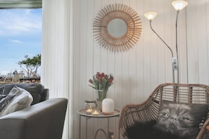 Una habitación con una silla de mimbre, un espejo redondo, un jarrón con flores y una mesa con velas.