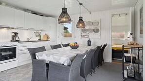 A modern kitchen with white cabinets, a dining table with a white tablecloth, wicker chairs, and pendant lights. - Cozy summer house close to the beach (Assens)