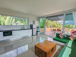 A modern kitchen with a large window, a dining area with a green sofa and a wooden coffee table, and a balcony with outdoor furniture. - Poolside Apartments and Rooms (Ko Chang)