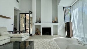 Een moderne woonkamer met een open haard, een bank, een salontafel en een groot raam met lichte gordijnen.