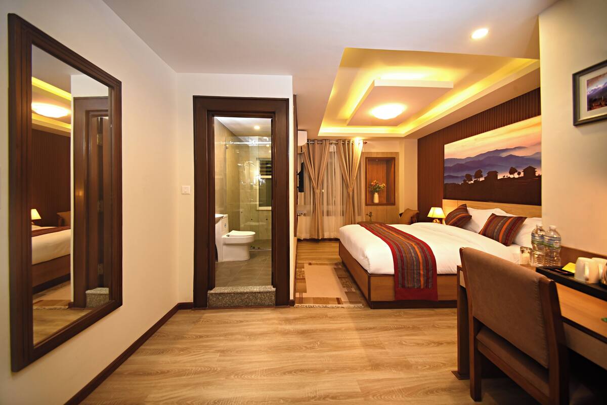 A hotel room with a large bed, a desk, a chair, a mirror, and a bathroom visible through an open door.