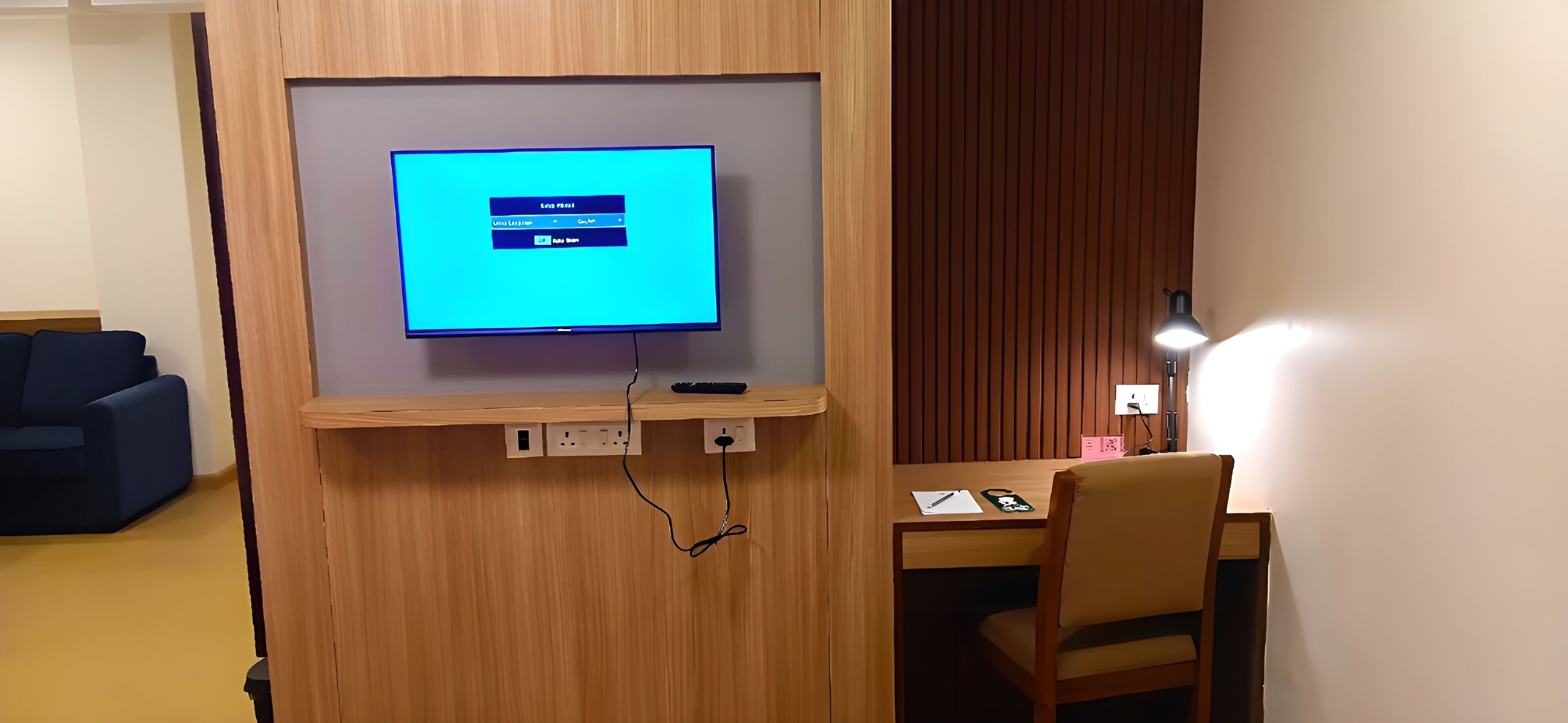 Una habitación con televisor montado en un panel de madera, un escritorio con una silla, una lámpara y un sofá.