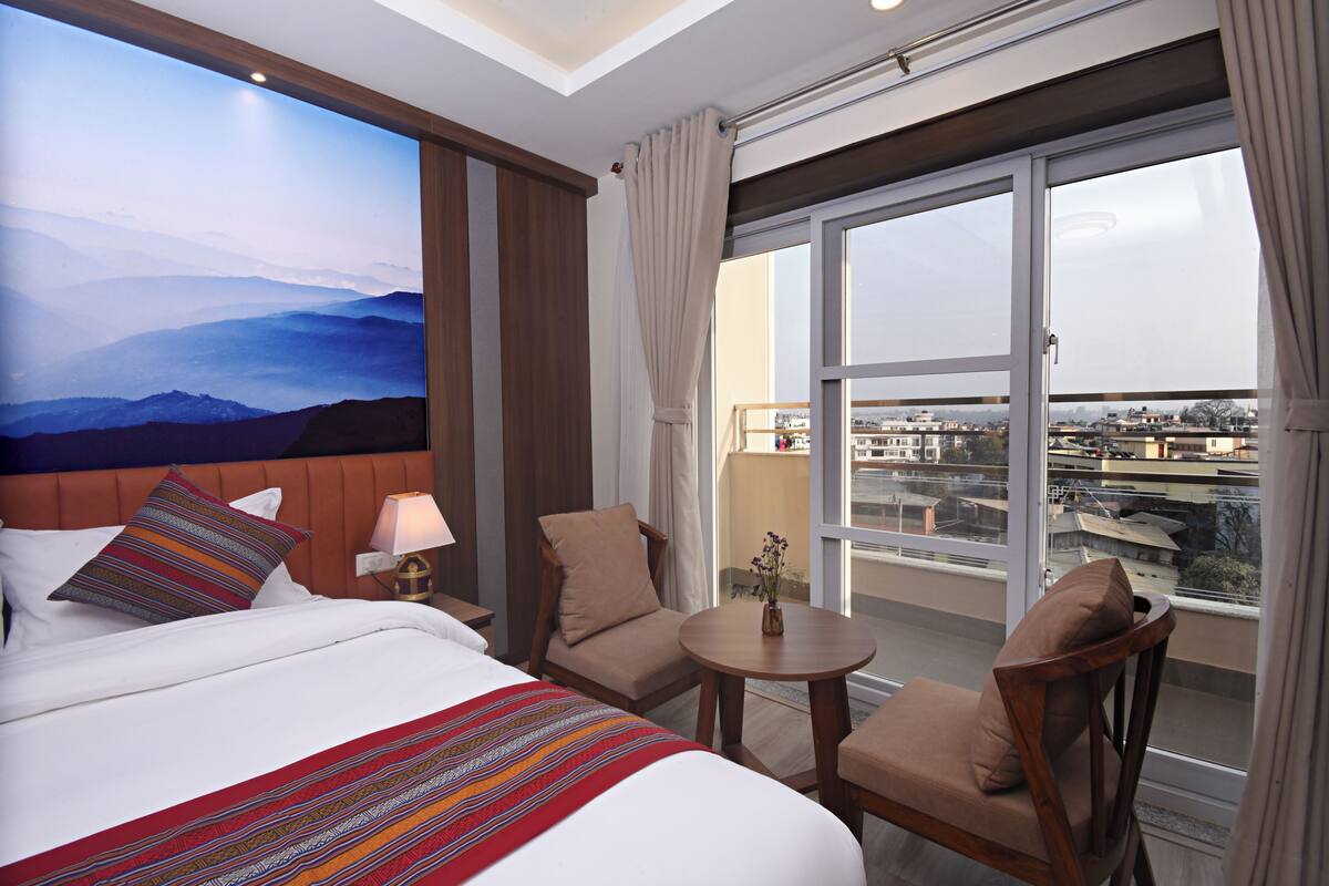 A hotel room with a large bed, two chairs, a small table, and a view of the city through large windows.