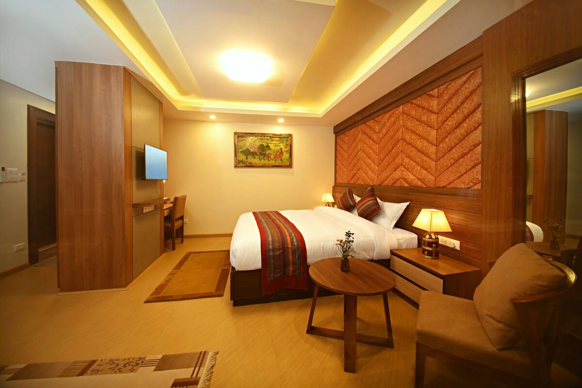 A hotel room with a bed, bedside tables, a TV, a wooden headboard, a painting, and a small table with a lamp.