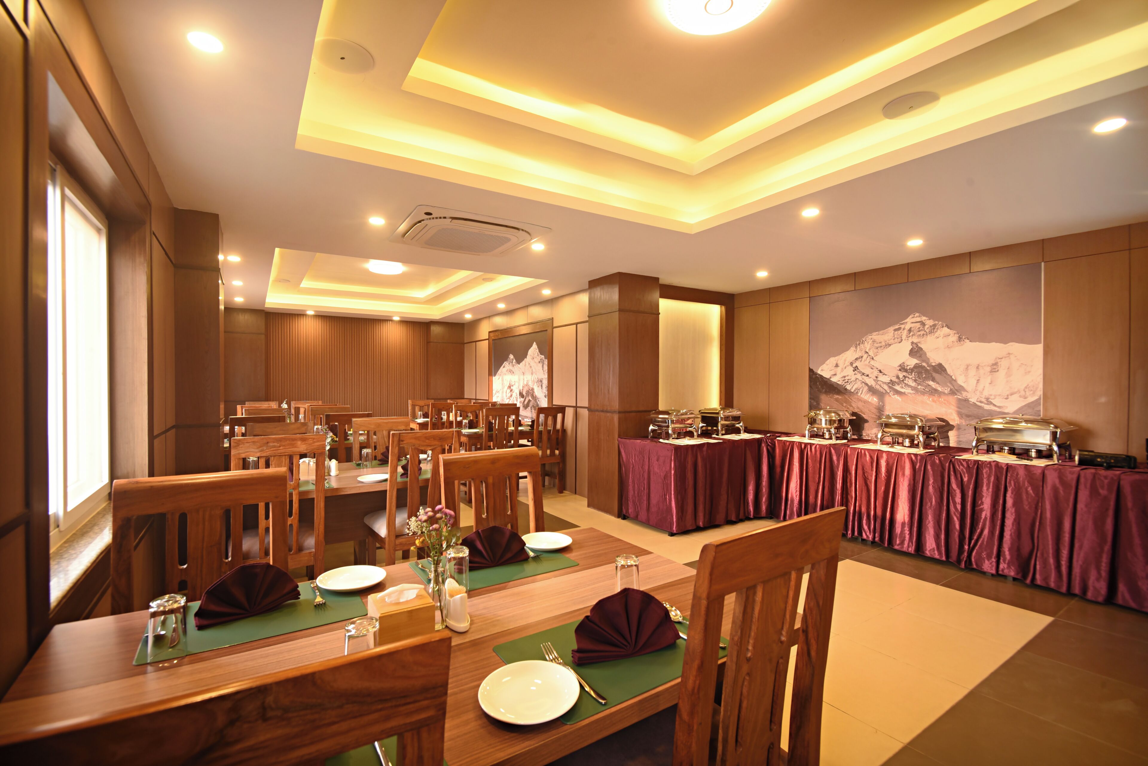 Un comedor con mesas y sillas de madera, una mesa de buffet con alimentos, y un mural grande en la pared.