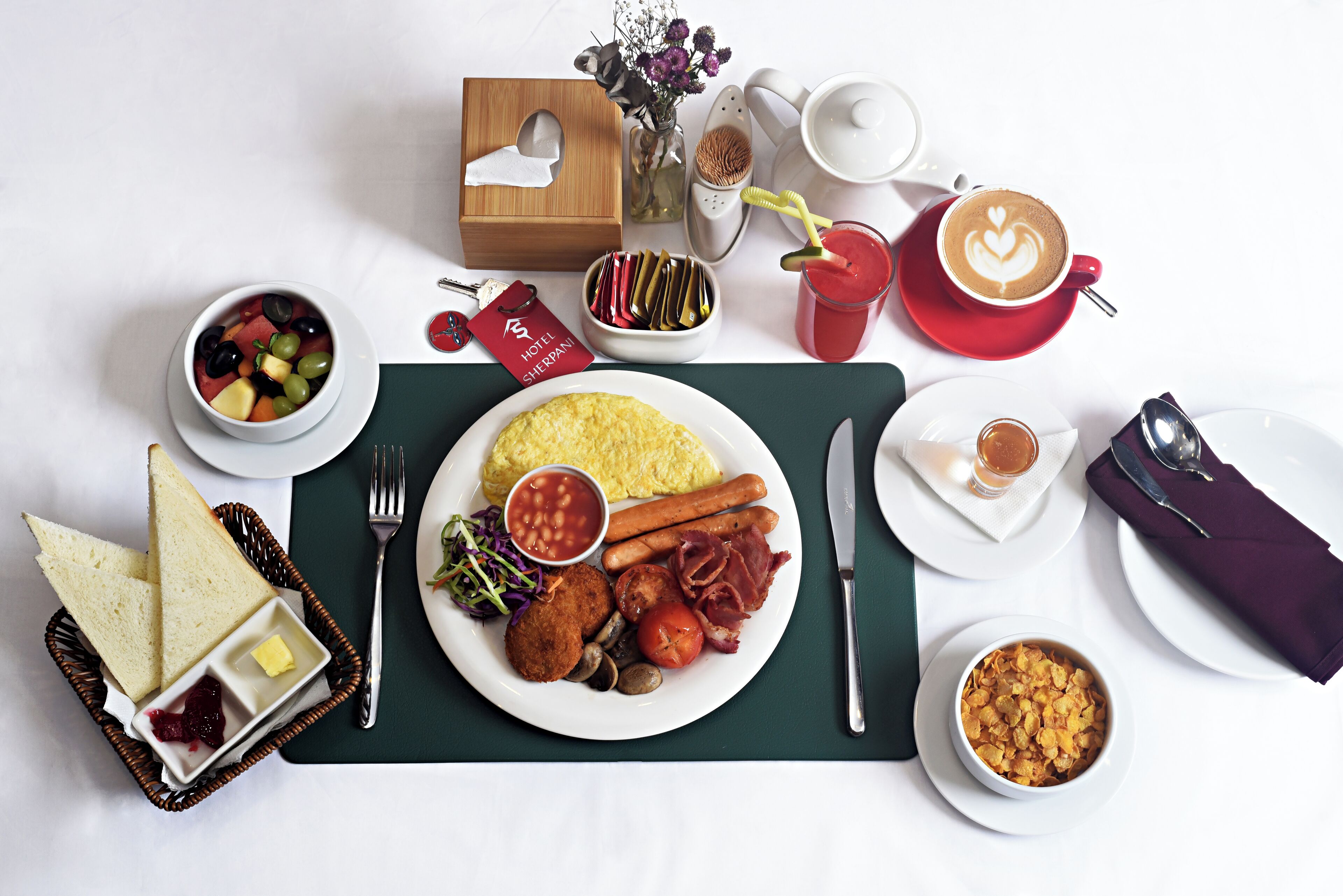 A breakfast setting with a plate of eggs, bacon, tomatoes, and mushrooms, a bowl of fruit, a cup of coffee, and a basket of bread.