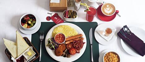 A breakfast setting with a plate of eggs, bacon, tomatoes, and mushrooms, a bowl of fruit, a cup of coffee, and a basket of bread.