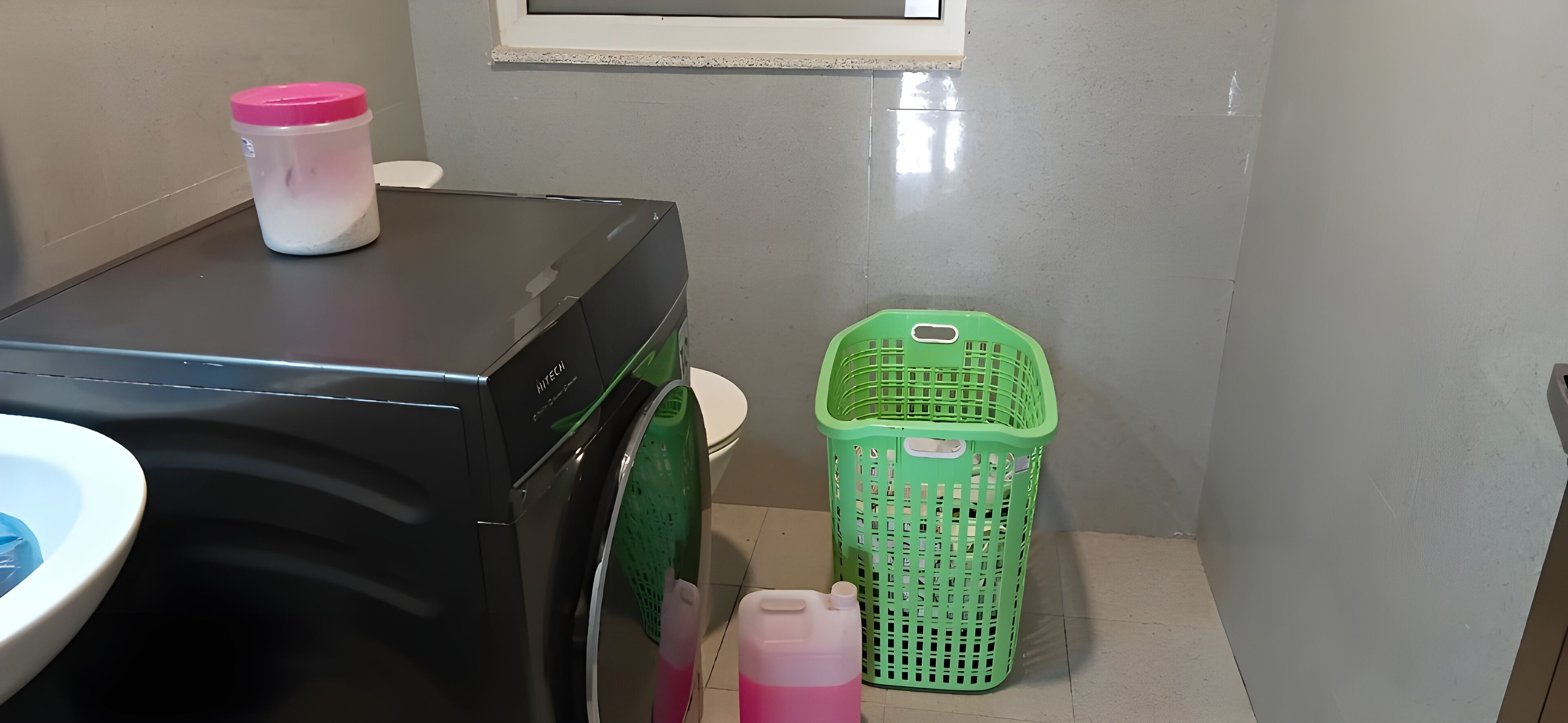 A compact bathroom with a washing machine, a green laundry basket, and a pink container.