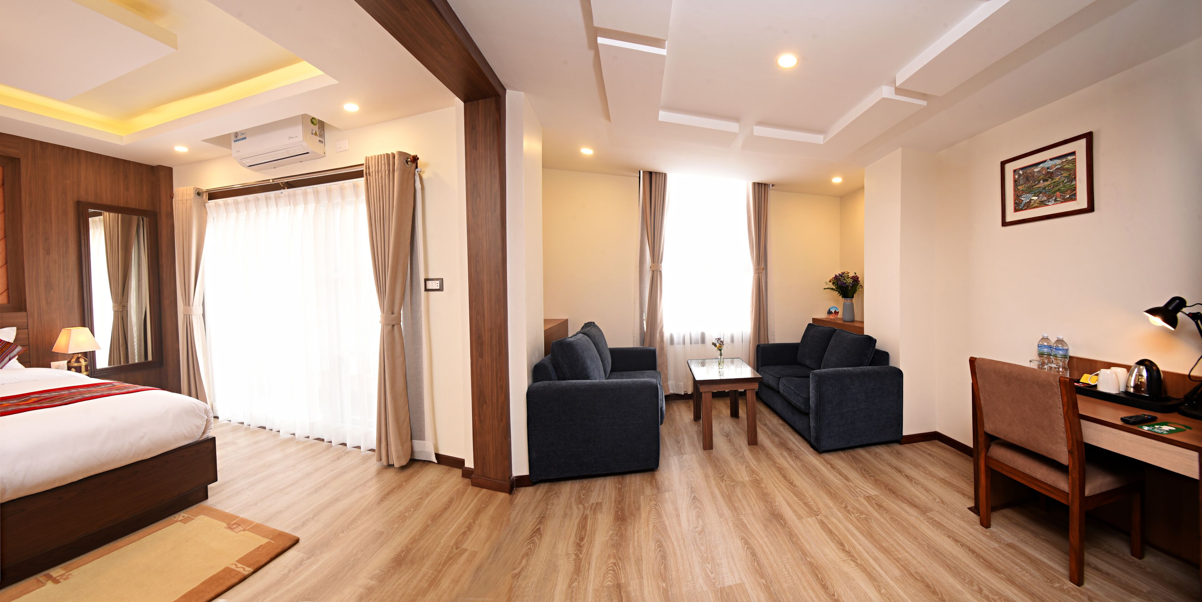 A hotel room with a bed, two sofas, a small table, a desk with a chair, and an air conditioning unit.