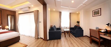 A hotel room with a bed, two sofas, a small table, a desk with a chair, and an air conditioning unit.