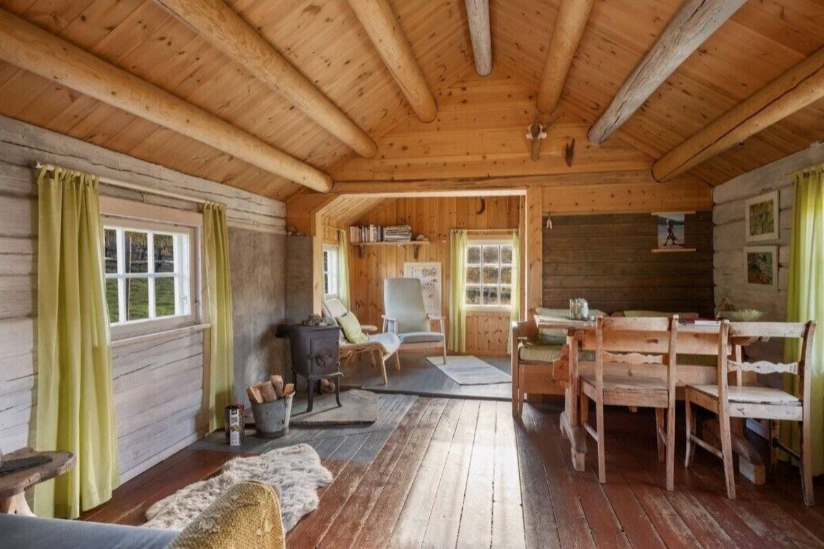 A cozy cabin interior with wooden walls and ceiling, a fireplace, a dining area with wooden chairs, and a window with curtains.