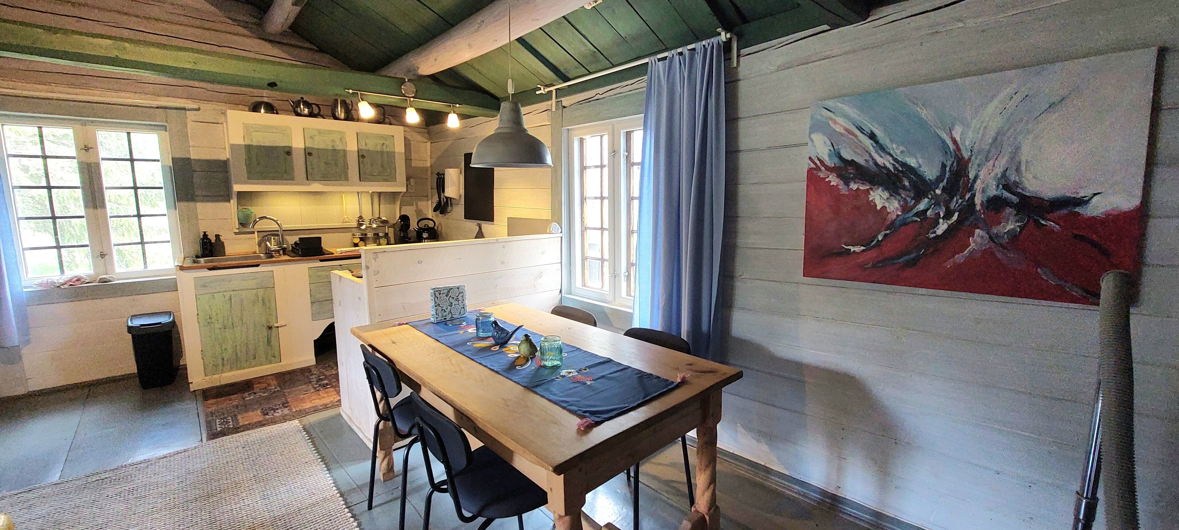 A rustic kitchen with wooden cabinets, a dining table with chairs, and a large abstract painting on the wall.