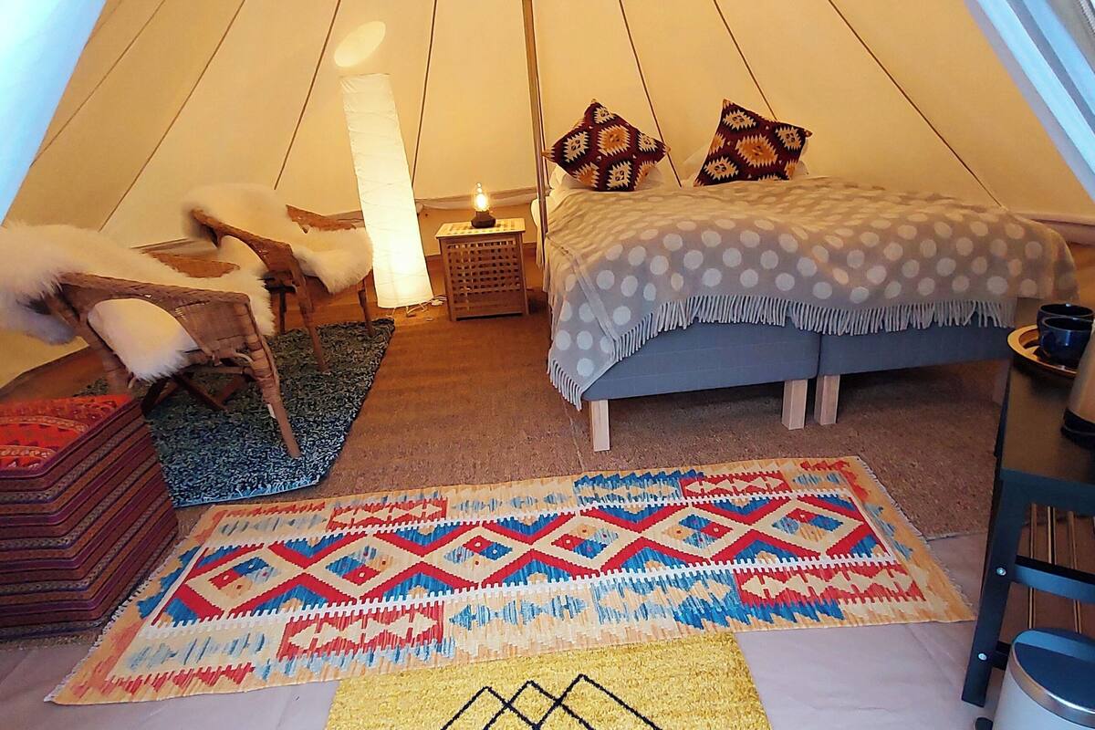 A cozy tented accommodation with a bed, a chair, a small table with a lamp, and a patterned rug.