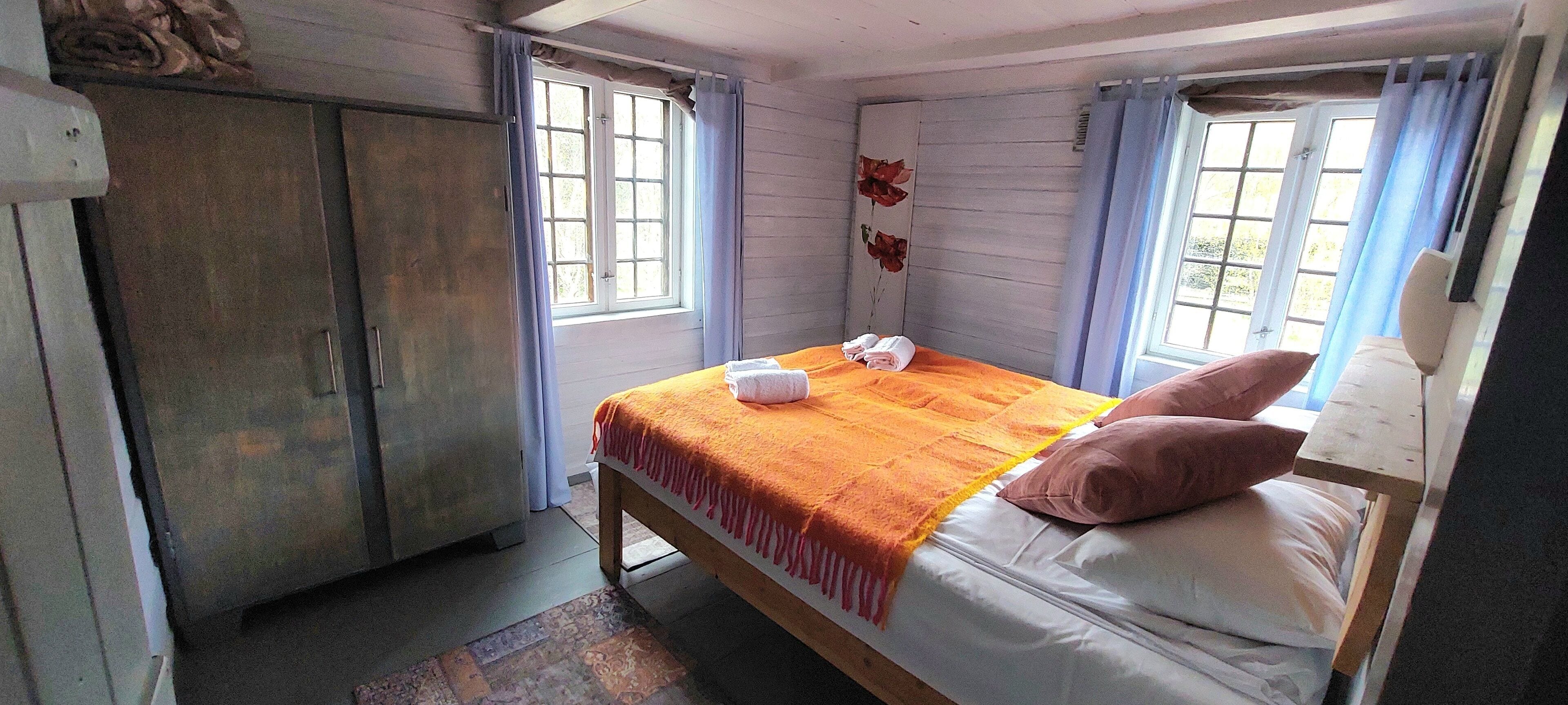 Un dormitorio con cama, almohadas, manta, armario y ventana con cortinas.