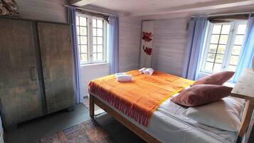 Un dormitorio con cama, almohadas, manta, armario y ventana con cortinas.