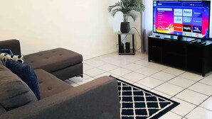 A living room with a sofa, a television displaying streaming services, a ceiling fan, and a cityscape painting on the wall. - 3-bedroom house in beautiful Eagle Pass with WiFi (Eagle Pass)