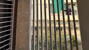 Un balcón con barandillas verticales, una ventana con persianas horizontales y vistas a un edificio con ropa tendida.