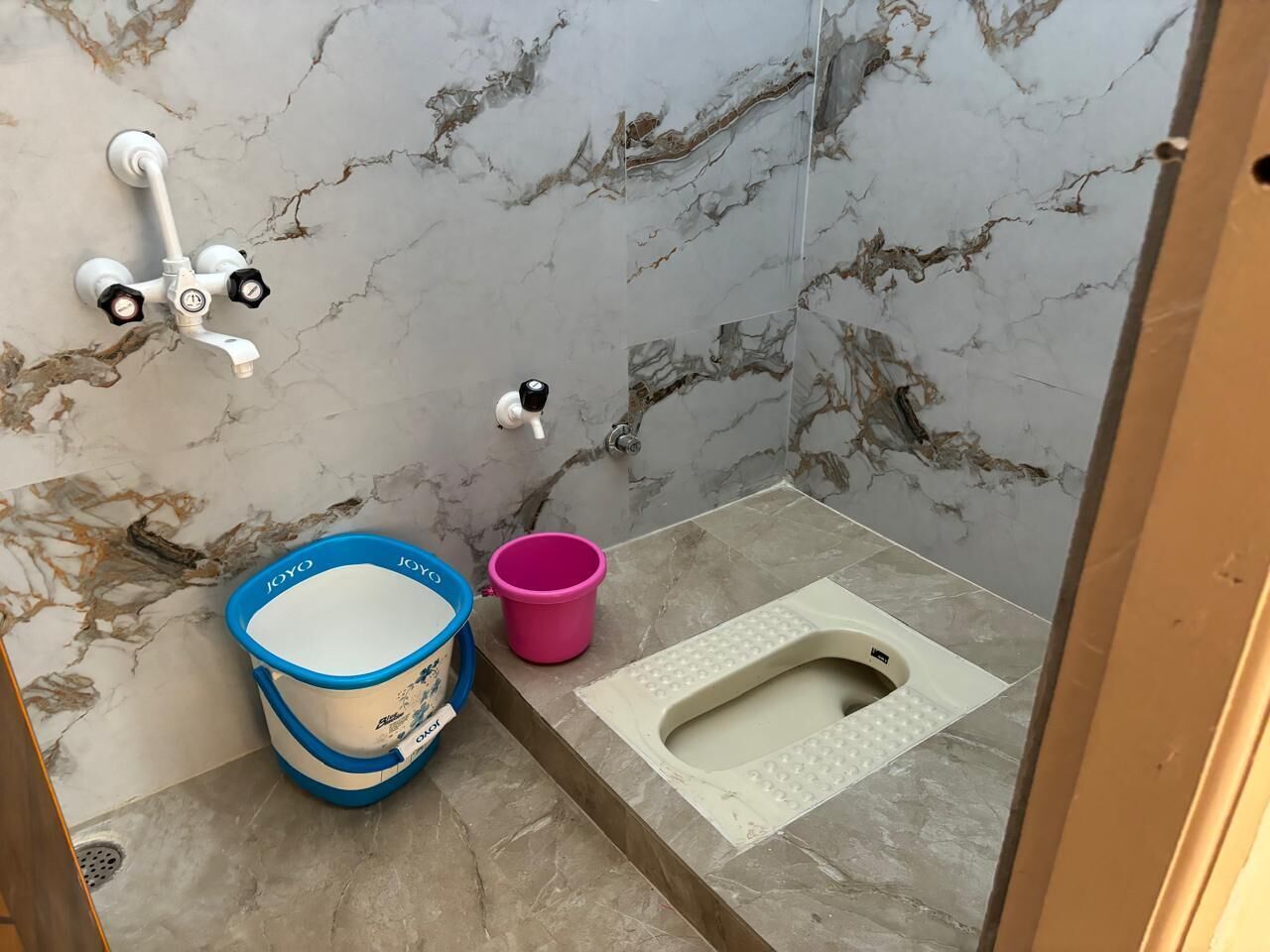 A bathroom with a marble wall, a white toilet, a blue and white bucket, and a pink bucket.