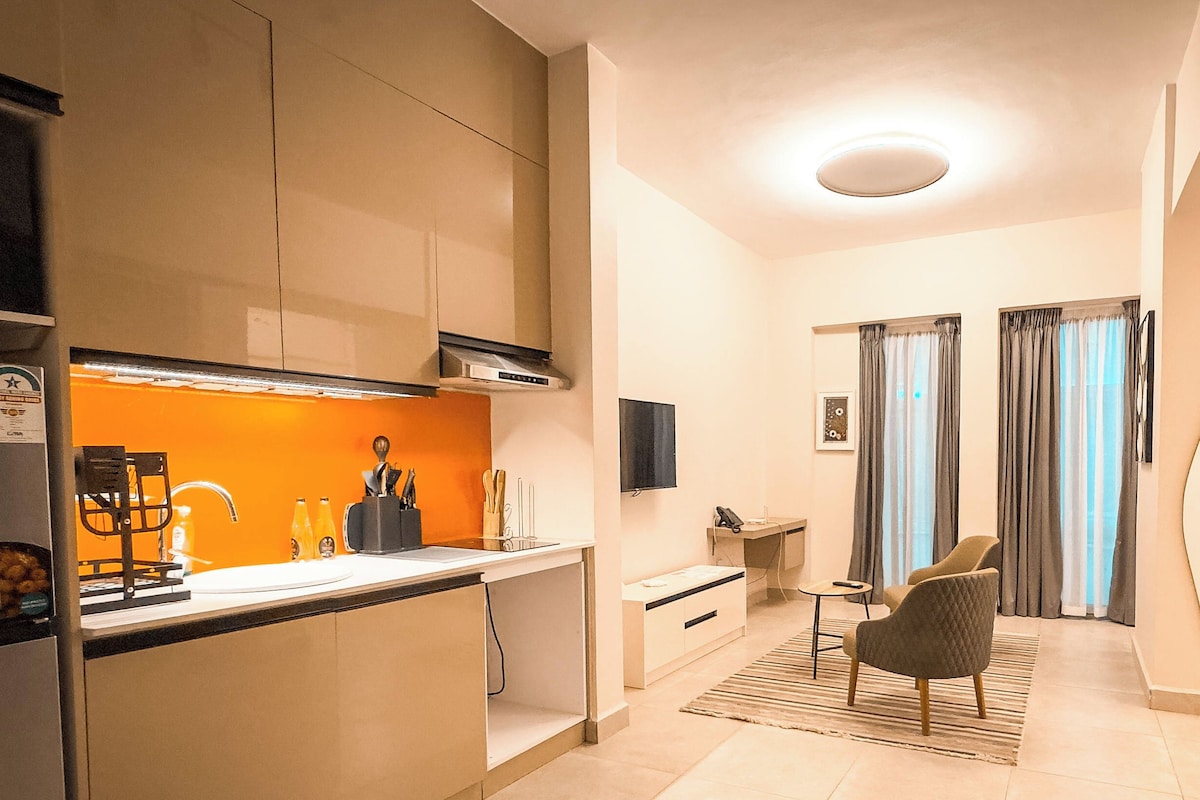 A modern kitchen with a built-in oven, a sink, and a countertop with kitchen utensils. There is a dining area with a table and chairs, and a small desk with a chair. The room has a neutral color scheme with beige walls and a light-colored floor.