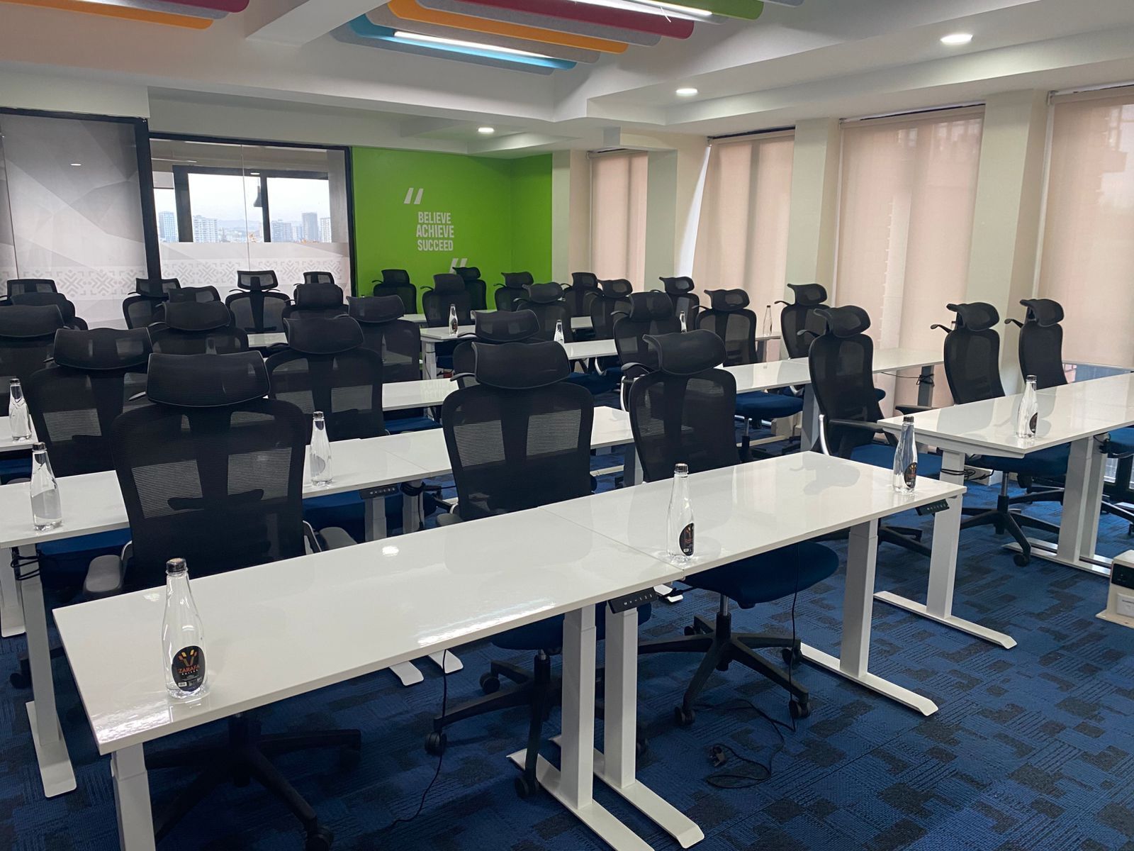 Una sala de conferencias con filas de sillas de oficina negras y mesas blancas, una cita motivacional en color verde en la pared y ventanas amplias con persianas.