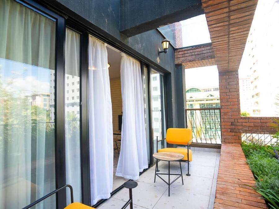 Un balcón con dos sillas amarillas y una mesita pequeña, ventanas amplias con cortinas blancas y un muro de ladrillo.