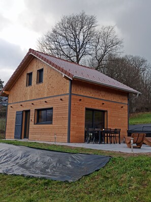 Ein modernes Holzhaus mit geneigtem Dach, großen Fenstern und einer Terrasse mit Gartenmöbeln.