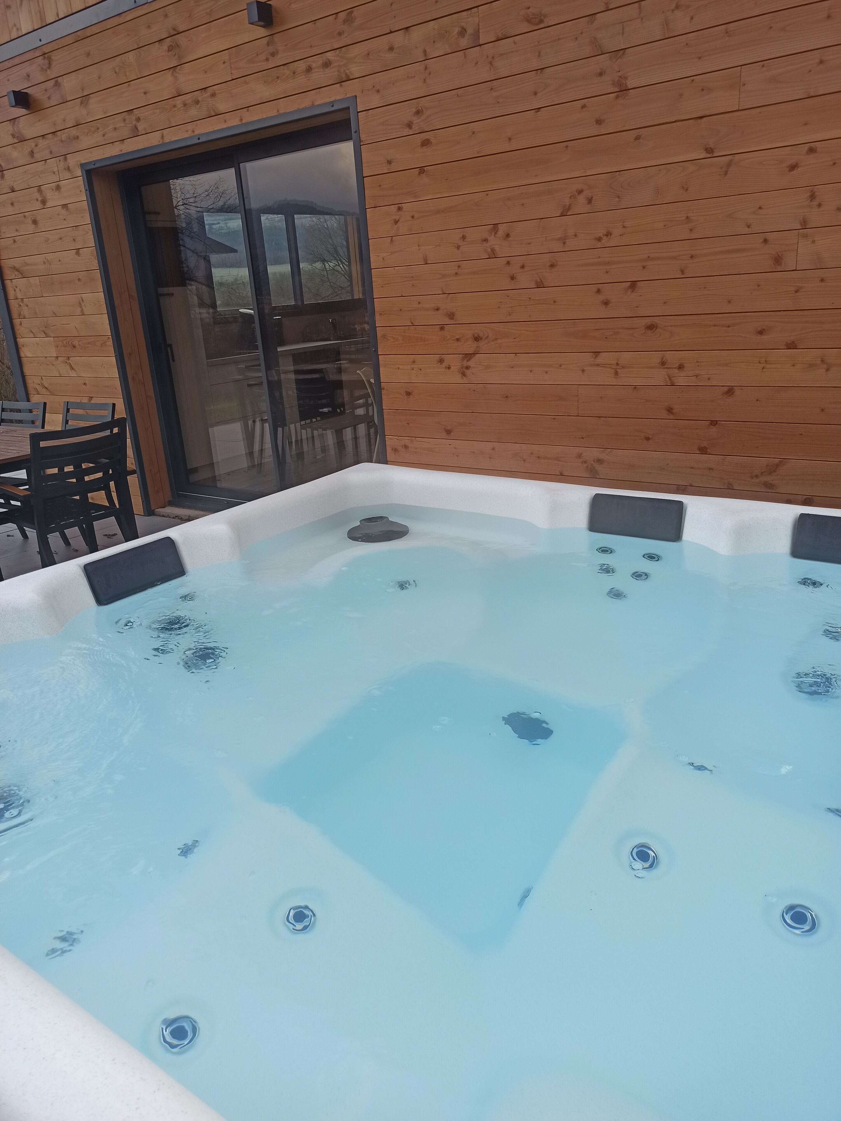 A hot tub with a wooden exterior wall and a glass door leading to an outdoor seating area.