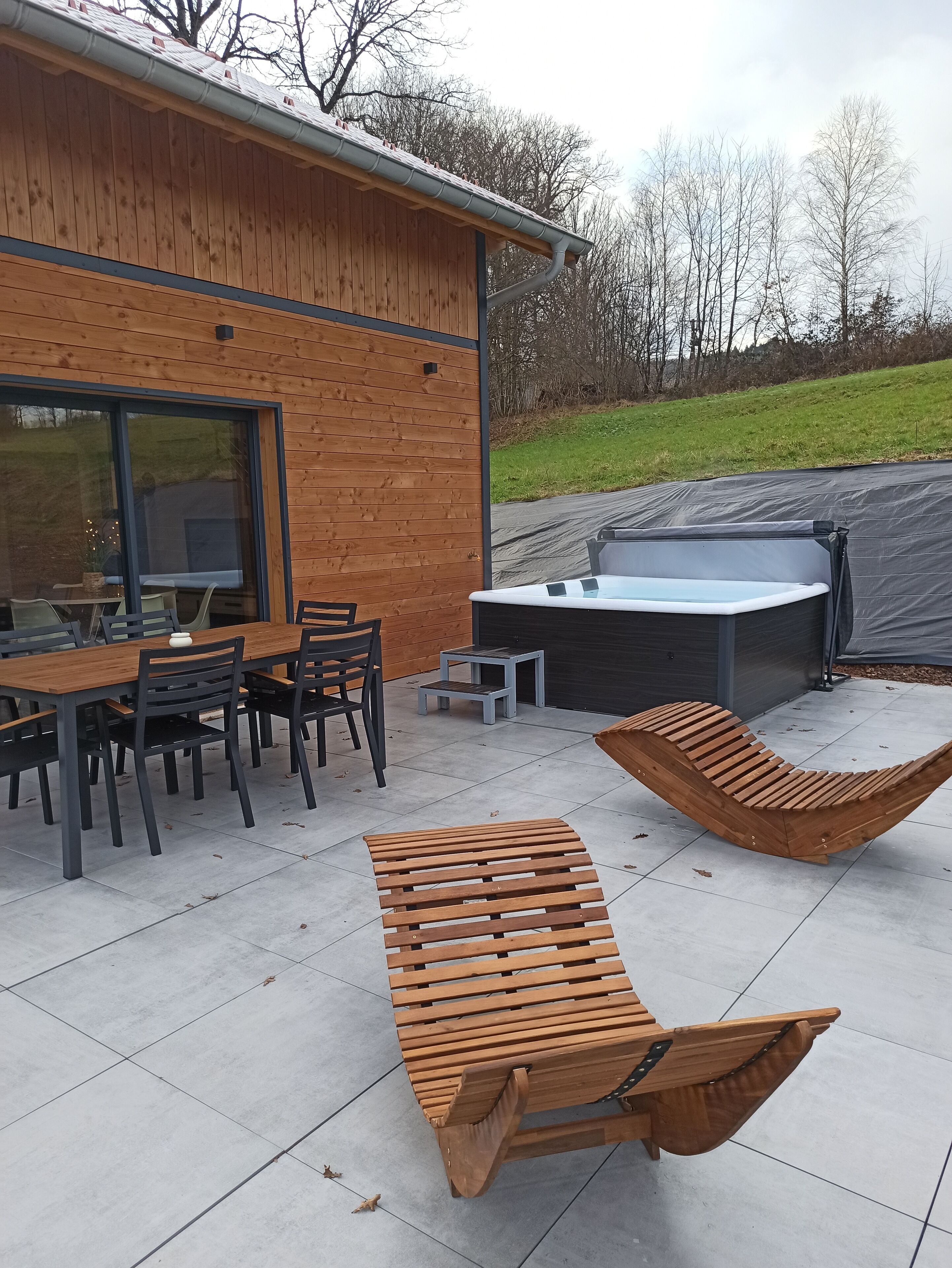 Un patio moderno al aire libre con mesa y sillas de madera, un jacuzzi y dos sillones de madera.