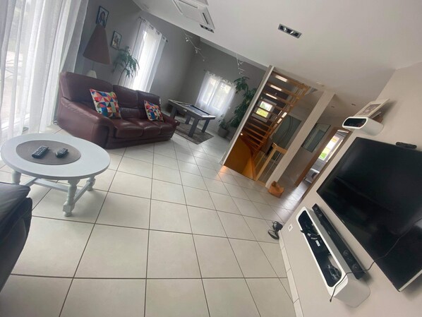 Uma sala de estar com um sofá marrom, uma mesinha redonda, uma televisão e uma escada que leva a outro andar.