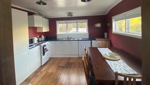 Een keuken met houten vloeren, witte kastjes, een eettafel met stoelen en een raam met uitzicht op bomen.