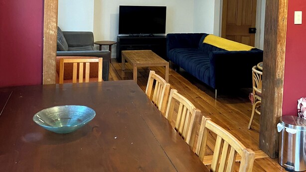 Een eethoek met een houten tafel en stoelen, een televisie op de achtergrond en een bank met een gele deken.