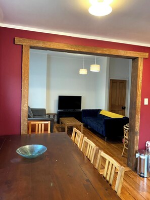 Un comedor con una mesa y sillas de madera, un televisor al fondo y un sofá con una manta amarilla.