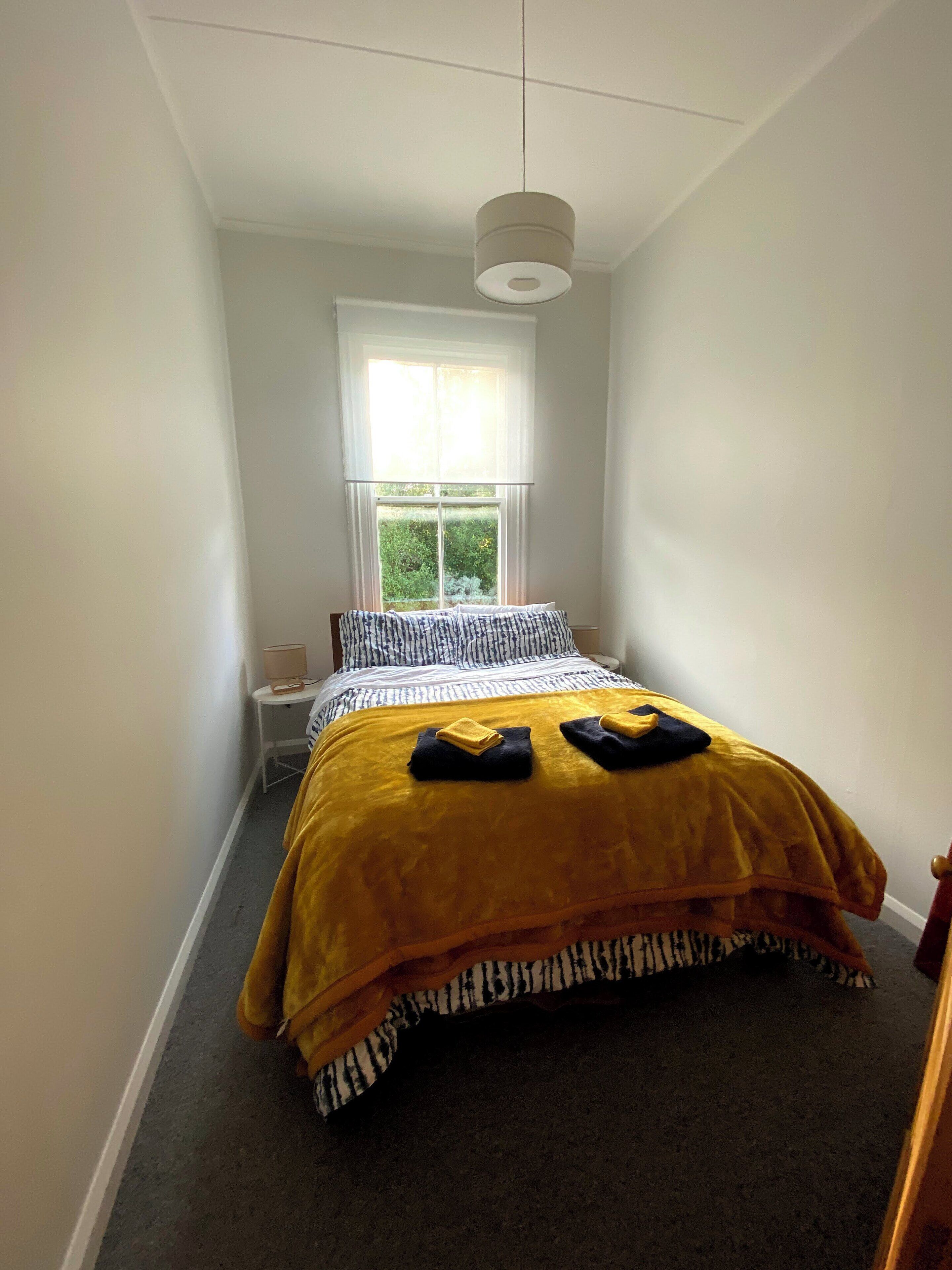 Une petite chambre à coucher, bien rangée, avec un lit, une table de chevet, une lampe et une fenêtre donnant sur un paysage verdoyant.