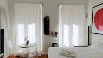 Una camera d'albergo con un letto, un tavolino con una sedia, un termosifone e un'ampia finestra con tende.