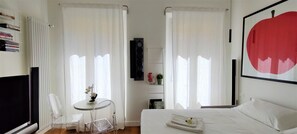 A hotel room with a bed, a small table with a chair, a radiator, and a large window with curtains. - NOVELLA STYLE FLORENCE 2 (Firenze)