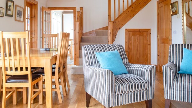 Een ruime woonkamer met een houten eettafel en stoelen, een gestreept fauteuil met een blauw kussen, en een houten trap.