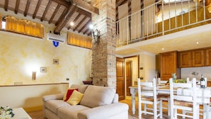 A spacious living area with a high ceiling, a stone pillar, a sofa with cushions, a dining table with chairs, and a kitchen in the background.