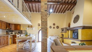 A spacious living area with a high ceiling, exposed wooden beams, a stone pillar, a fireplace, and a dining area with a table and chairs.
