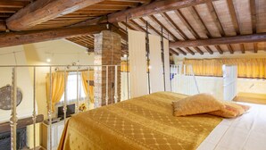 A bedroom with a bed, wooden beams, a brick fireplace, and a window with curtains.