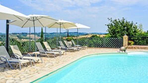 A poolside area with lounge chairs and umbrellas, a stone-tiled deck, and a view of the countryside.