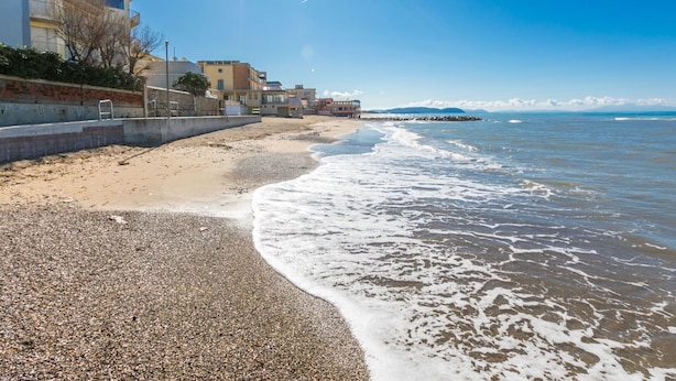 A beach with pebbly shore, a clear sky, and buildings in the background.