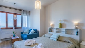 A bedroom with a bed, a blue chair, a bedside table with a lamp, a shelf with books, and a window with curtains.
