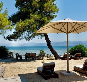 A beachside lounge area with sun loungers and a large umbrella, overlooking a calm sea and distant mountains. - SunBreeze-Beachfront Luxury Villa 5 Bed/4 bath- Sleeps 10 (Lefkimmi)