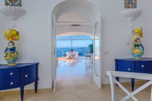 An open doorway leading to a balcony with a view of the sea, a white chair, and two blue side tables with decorative vases. - Estate4home - Regina Dar (Positano)