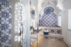 A bathroom with a glass shower enclosure, a sink, and a decorative tiled wall. - Estate4home - Regina Dar (Positano)