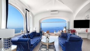 Amplia sala de estar con un gran ventanal arqueado que ofrece vista al mar, amueblada con sofás azules y una mesa de centro.